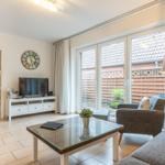 Wohnzimmer mit Glas-Tisch, Sofa und Sessel. Durchgang zur Küche. Fenster zur Terrasse.