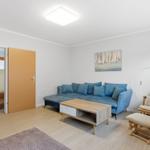 Wohnzimmer mit blauem Sofa, Holztisch und Schaukelstuhl. An der Wand hängt ein Bild.