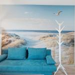 Ein blauer Sofa mit Kissen vor einer Wandtapete mit Strandmotiv und einem weißen Kleiderständer.