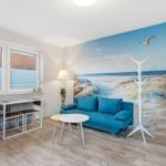 Wohnzimmer mit blauem Sofa, Wandbild Strand und Fenster mit Jalousien.