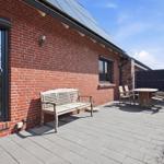 Terrasse mit Holzbank, Tisch und Stühlen neben rotem Backsteinhaus unter blauem Himmel.