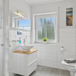 Badezimmer mit Dusche, Waschtisch und Toilette. Fenster mit Pflanze und Wandbild.