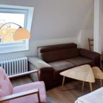 Wohnzimmer mit braunem Sofa, pinkem Sessel und Holztisch. Fenster mit Blick auf Dächer.