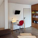 Schreibtisch mit roter Drehstuhl, Wandfernseher und Holzregal mit Büchern und Spielzeug.