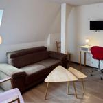 Ein Wohnraum mit braunem Sofa, Holztisch und roter Drehstuhl. An der Wand hängt ein Fernseher.