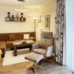 Ein Wohnzimmer mit braunem Sofa, grauer Sessel und Glas-Tisch. Vorhänge und Wandbild sind sichtbar.