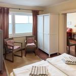 Zimmer mit Bett, zwei Stühlen, Schrank und Fenster. Durchgang zur Wohnfläche mit Kamin.