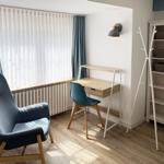 Ein Zimmer mit Schreibtisch, Stuhl, Kleiderständer und Fenster mit Vorhängen.