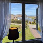 Durch das Fenster blickt man auf eine grüne Wiese, Häuser und das Meer.