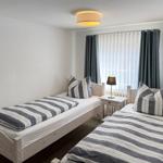 Zwei Einzelbetten mit gestreiften Bettwäsche, Nachttisch mit Lampe und Fenster mit Vorhängen.
