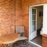 Terrasse mit Holztisch und Stuhl, rote Ziegelwand, Tür zum Innenraum.
