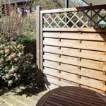 Holzterrasse mit Tisch, Sitzgelegenheit und Pflanzen. Holzgeländer mit Gitter oberhalb.