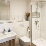 Badezimmer mit Dusche, Waschbecken und Toilette. Weiße Fliesen und Holzunterschrank.