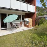 Terrasse mit Tisch, Stühlen und Sonnenschirm. Strandkorb und Grünfläche im Hintergrund.