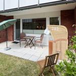 Terrasse mit Tisch, zwei Stühlen und Sonnenschirm. Holzstuhl und Korb neben grüner Pflanze.