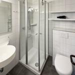 Badezimmer mit Dusche, Waschbecken und WC. Weiße Fliesen und graue Bodenfliesen.