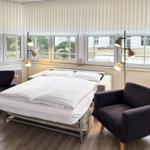 Schlafzimmer mit Bett, zwei Sesseln und Fenstern mit Jalousien.