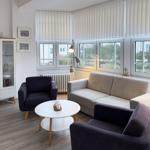 Wohnzimmer mit Sofa, zwei Sesseln, Tisch und Fenster. Regal und Lampen sind vorhanden.