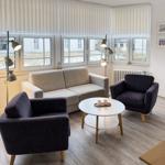 Wohnzimmer mit Sofa, zwei Sesseln und runden Tisch. Fenster mit Jalousien und Kühlschrank.