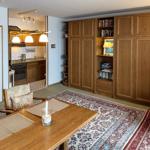 Dining area with wooden table, chairs, and rug. Kitchen and bookshelves are visible.