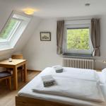 Schlafzimmer mit Doppelbett, Tisch, zwei Stühlen und Fenstern mit Blick auf Grün.