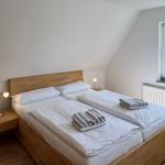 Zimmer mit Doppelbett, Holzbett, Nachttisch und Fenster mit Blick auf Bäume.