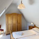 Schlafzimmer mit Dachschrägen, Holzschrank, Bett und Stuhl. Fenster und Lampen sichtbar.