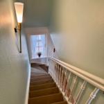 Treppe mit Teppich, weißer Handlauf und Wandlampe. Fenster mit Vorhängen im oberen Bereich.