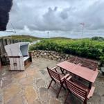 Terrasse mit Tisch, Stühlen und Liege. Steinplatten, Steinmauer, Grünflächen und Flagge.