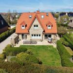 Haus mit rotem Dach, Garten und Terrasse. Umgeben von grüner Vegetation und Nachbarhäusern.