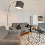 Wohnbereich mit grauem Sofa, zwei Sesseln und einem Tisch mit Wein. Hintergrund: Küchenzeile und Tür.