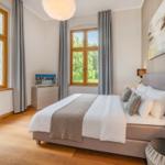 Schlafzimmer mit großem Bett, Holzboden und zwei Fenstern mit Blick auf Grün.