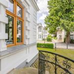 Haus mit weißer Fassade, Holzfenster und schmiedeeisernem Geländer. Grünanlage und Bäume im Hintergrund.