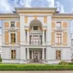Elegantes Haus mit goldverzierten Fenstern und Balkon. Grünflächen vor dem Eingang.