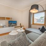 Wohnzimmer mit Sofa, TV und Fensterblick. Es gibt einen Tisch und eine Stehlampe.