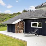 Schwarzes Haus mit Holzstapel, Liegestuhl und Terrasse vor grüner Landschaft.