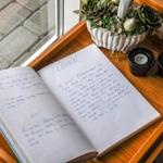 Ein Buch mit handschriftlichen Einträgen liegt auf einem Holztisch neben einer Pflanze und Kerzen.