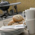 Tisch mit Brot, Tassen und Geschirr vor Grill und gelbem Mauerwerk.