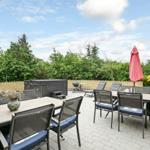 Terrasse mit Tisch, Stühlen, Sonnenschirm und Grill. Hintergrund: Grünflächen und Hecke.