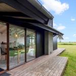 Modern villa with wooden terrace and large glass area. View of green field and trees.