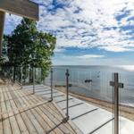 Terrasse mit Holz- und Betonfläche, Glasgeländer und Blick auf See und Strand.
