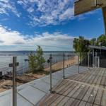 Terrasse mit Holz- und Glasgeländer überblickt einen Strand und See unter blauem Himmel.