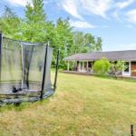 House with terrace, garden, and trampoline. Surrounded by trees under blue sky.