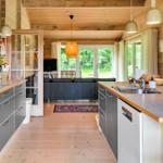 Kitchen with countertop, sink, and cabinets. In the background, living room with sofa and windows.