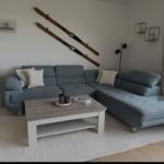 Ein Wohnzimmer mit grauem Sofa, Holztisch und Skiern an der Wand.