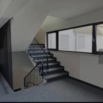 Treppe mit Geländer führt zu oberen Etagen. Große Fenster bieten Blick auf Außenbereich.