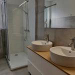 Badezimmer mit Dusche, Toilette und zwei Waschbecken auf Holzplatte.