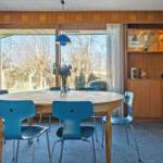 Esszimmer mit runden Tisch, blauen Stühlen und Holzwandverkleidung. Große Fenster mit Blick auf Außenbereich.