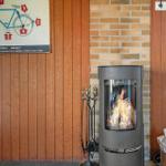Kamin mit brennendem Holz, daneben Feuerzeug und Holzstapel. An der Wand hängt ein Fahrrad-Info-Plakat.