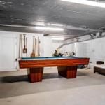 Pool table with blue felt and wooden frame in basement. Tools hang on the wall. A seating area stands beside it.